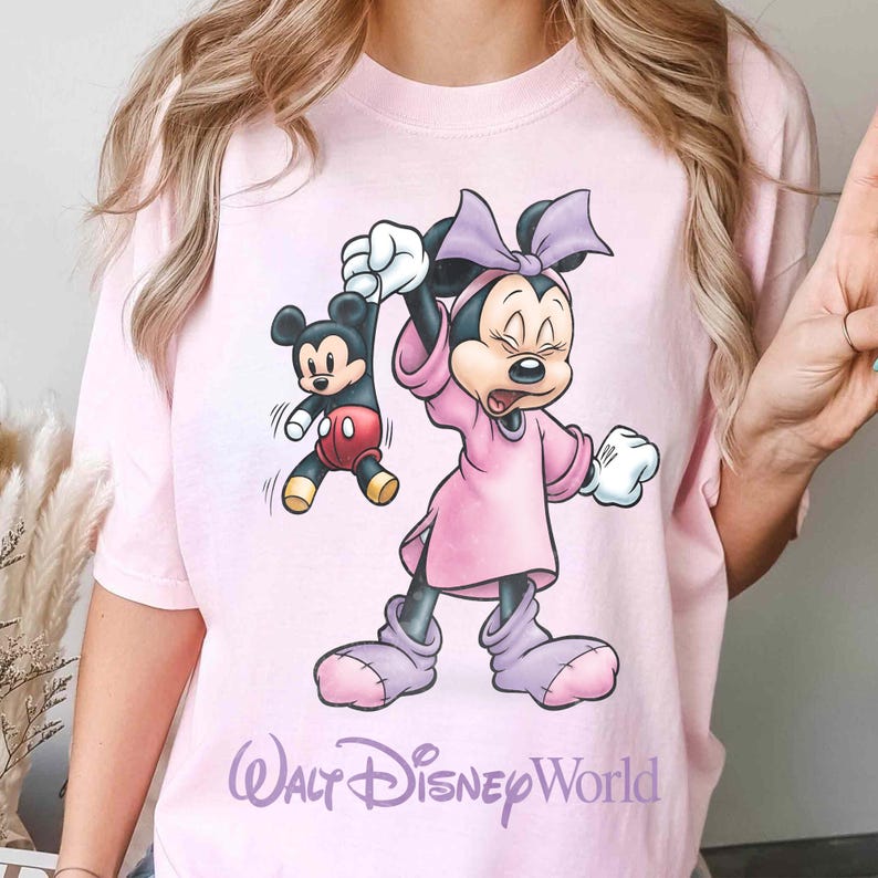 May include: A light pink t-shirt featuring a cartoon Minnie Mouse character holding a Mickey Mouse character. Minnie is wearing a pink dress and bow. The text "Walt Disney World" is printed in a cursive font at the bottom of the shirt.