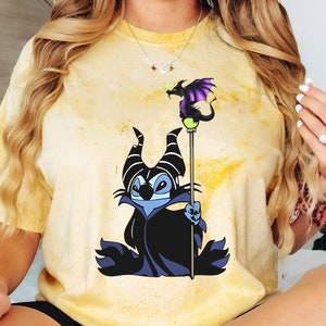 May include: A yellow tie-dye t-shirt with a cartoon image of Maleficent from Disney's Sleeping Beauty. Maleficent is depicted as a Stitch-like character with purple horns and a black cape. She is holding a staff with a purple dragon on top.