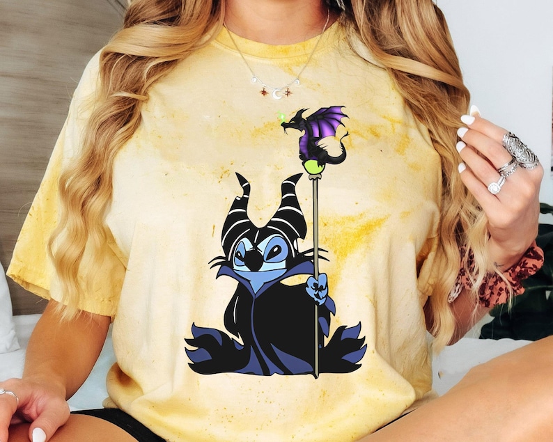 May include: A yellow tie-dye t-shirt with a cartoon image of Maleficent from Disney's Sleeping Beauty. Maleficent is depicted as a Stitch-like character with purple horns and a black cape. She is holding a staff with a purple dragon on top.