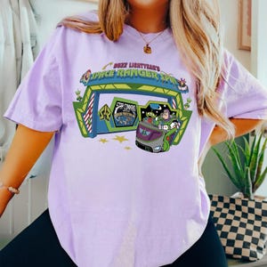 K&ouml;nnte beinhalten: Helllila T-Shirt mit einer Grafik von Buzz Lightyear's Space Ranger Spin. Das Design zeigt den Eingang der Attraktion, Buzz Lightyear und andere Charaktere. Das T-Shirt hat kurze &Auml;rmel.