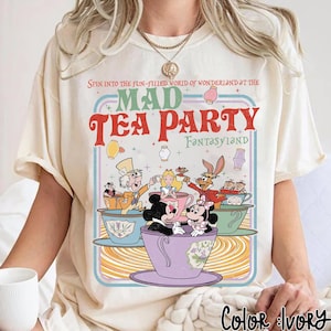 Puede incluir: Camiseta color marfil con un dise&ntilde;o colorido de Mad Tea Party, con Mickey y Minnie Mouse, Alicia y el Sombrerero Loco. El texto dice: "Spin into the fun-filled world of Wonderland at the Mad Tea Party Fantasyland."