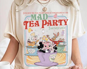 May include: Ivory-colored t-shirt featuring a colorful Mad Tea Party design with characters like Mickey and Minnie Mouse, Alice, and the Mad Hatter. The text reads "Spin into the fun-filled world of Wonderland at the Mad Tea Party Fantasyland."