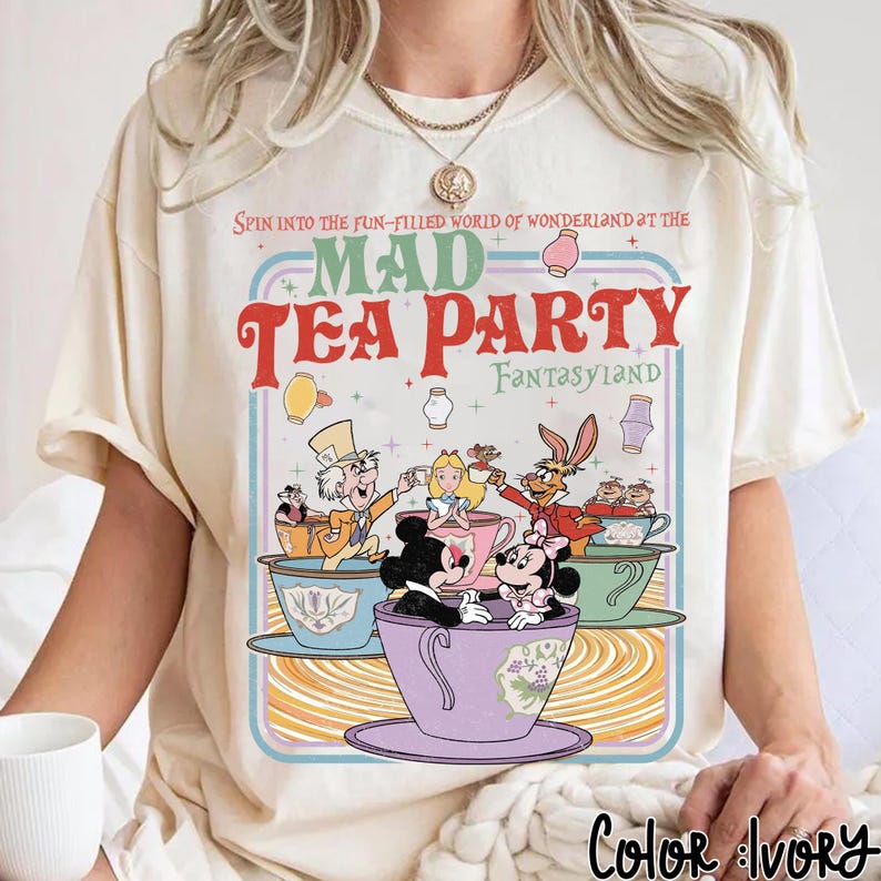 Puede incluir: Camiseta color marfil con un dise&ntilde;o colorido de Mad Tea Party, con Mickey y Minnie Mouse, Alicia y el Sombrerero Loco. El texto dice: "Spin into the fun-filled world of Wonderland at the Mad Tea Party Fantasyland."