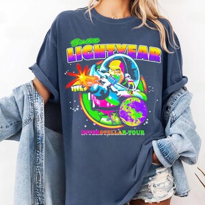 May include: A navy blue t-shirt featuring a colorful graphic of Buzz Lightyear in a space suit, with the text "Buzz Lightyear" above and "Interstellar Tour" below. The design includes a planet and a laser blast.