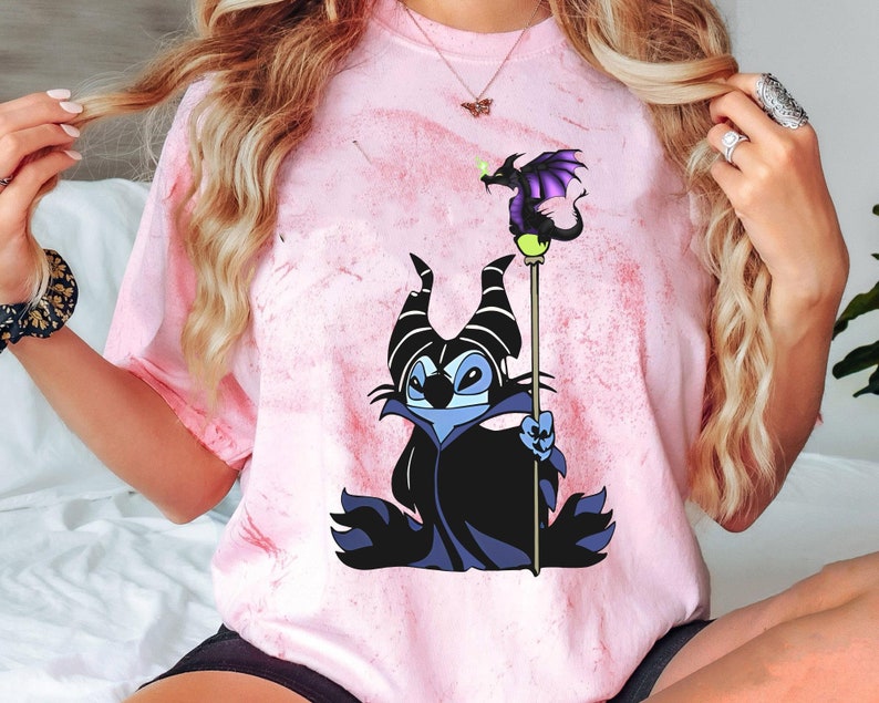 May include: A pink tie-dye t-shirt with a cartoon image of Stitch dressed as Maleficent from Disney's Sleeping Beauty. Stitch is holding a staff with a purple dragon on top.