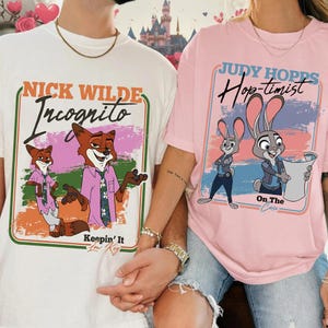May include: Two t-shirts, one white and one pink, featuring cartoon characters. The white shirt displays "Nick Wilde Incognito" with a fox illustration. The pink shirt shows "Judy Hopps Hop-timist" with a bunny illustration. Both shirts have colorful designs.