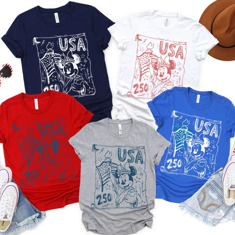 May include: A collection of five t-shirts in various colors: navy blue, white, red, gray, and blue. Each shirt features a graphic of Mickey Mouse holding a torch and American flag, with the word "USA" and the number "250" printed on a scroll.