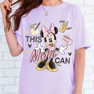Może przedstawiać: Jasnofioletowa koszulka z Minnie Mouse w r&oacute;żowej sukience i kokardzie. Koszulka ma napis "THIS MOM CAN", listę rzeczy do zrobienia, telefon i kubek. Projekt zawiera serca i logo Disneya.