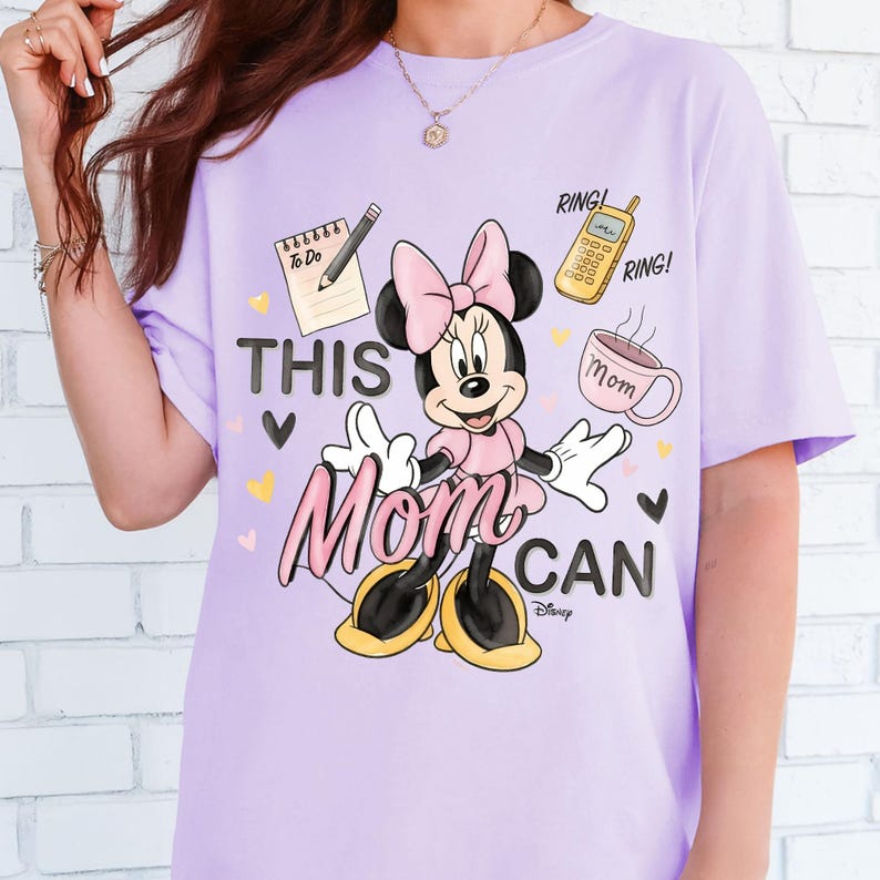 Może przedstawiać: Jasnofioletowa koszulka z Minnie Mouse w r&oacute;żowej sukience i kokardzie. Koszulka ma napis "THIS MOM CAN", listę rzeczy do zrobienia, telefon i kubek. Projekt zawiera serca i logo Disneya.