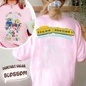 Pu&ograve; includere: T-shirt Comfort Colors rosa chiaro con una grafica di un treno a fumetti sul retro e una grafica a fumetti colorata sul davanti. La grafica frontale include il testo "WHERE ANYONE CAN BE ANYTHING". La maglietta &egrave; di colore rosa chiaro.