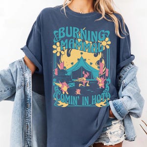 Puede incluir: Camiseta azul marino con un dise&ntilde;o gr&aacute;fico con el texto "BURNING MAMMAL COMIN' IN HOT". El dise&ntilde;o incluye una tienda de campa&ntilde;a, personajes de dibujos animados y llamas en amarillo, rosa y azul. La camiseta tiene mangas enrolladas.