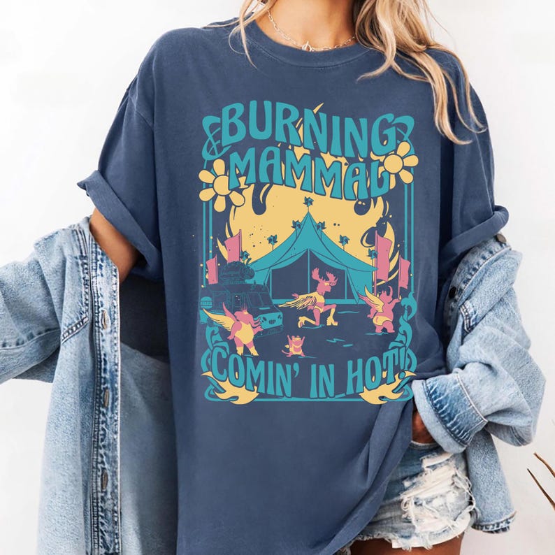 Puede incluir: Camiseta azul marino con un dise&ntilde;o gr&aacute;fico con el texto "BURNING MAMMAL COMIN' IN HOT". El dise&ntilde;o incluye una tienda de campa&ntilde;a, personajes de dibujos animados y llamas en amarillo, rosa y azul. La camiseta tiene mangas enrolladas.