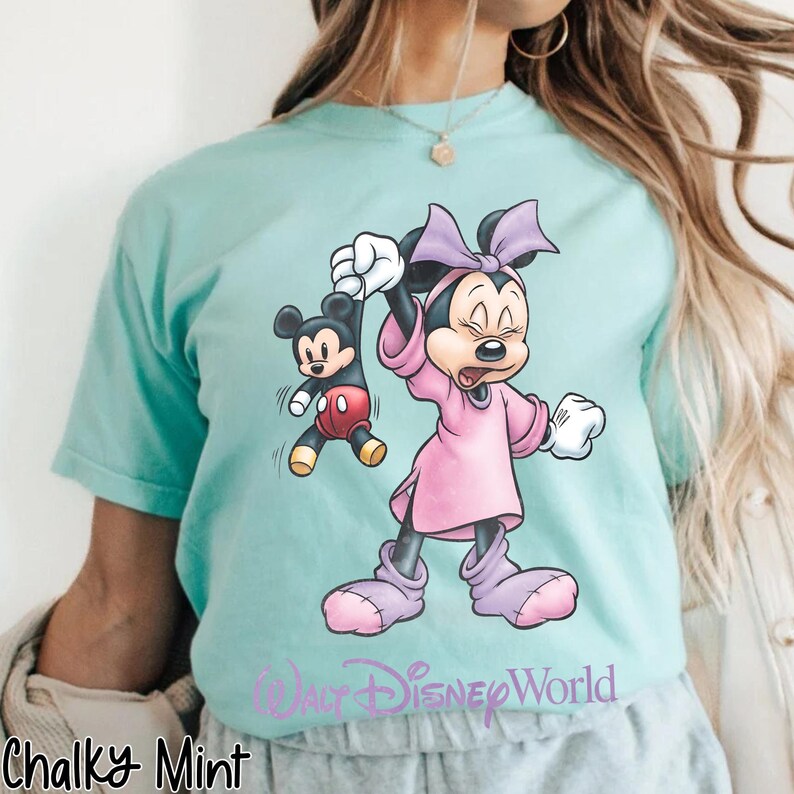May include: A light teal t-shirt featuring a cartoon Minnie Mouse holding Mickey Mouse. Minnie is wearing a pink dress and bow. The text "Walt Disney World" is below the characters. The shirt also has the text "Chalky Mint" at the bottom.