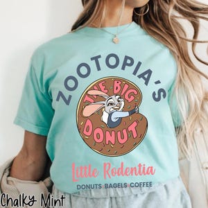 Puede incluir: Camiseta azul claro con una imagen de dibujos animados de Judy Hopps de Zootopia dentro de una rosquilla. El texto de la camiseta dice "Zootopia's The Big Donut" y "Little Rodentia, Donuts Bagels Coffee".