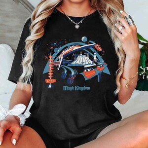 Op de afbeelding: Zwart t-shirt met een kleurrijke afbeelding van een themaparksc&egrave;ne met een monorail, een raket en de tekst "Magic Kingdom". Het ontwerp bevat ook een retro-bord met de tekst "Land".