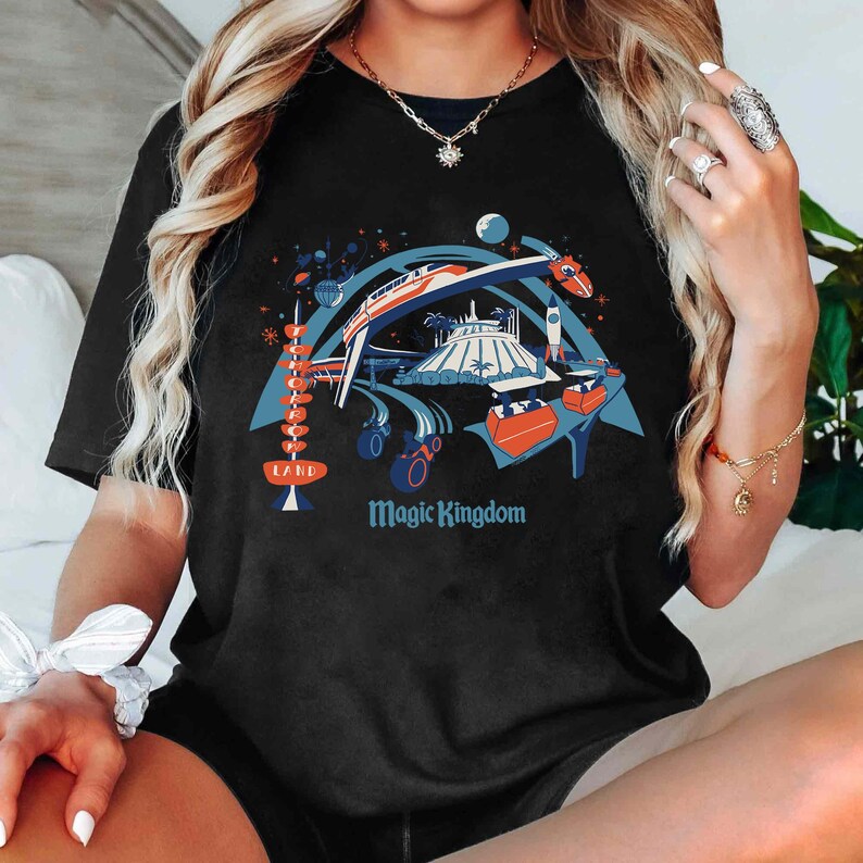 Op de afbeelding: Zwart t-shirt met een kleurrijke afbeelding van een themaparksc&egrave;ne met een monorail, een raket en de tekst "Magic Kingdom". Het ontwerp bevat ook een retro-bord met de tekst "Land".
