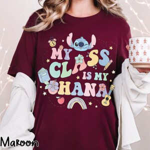 Puede incluir: Camiseta color granate con el texto "My Class is My Ohana" y un personaje de dibujos animados. El dise&ntilde;o incluye un reloj, un libro, flores, un l&aacute;piz, un ukelele y un arco&iacute;ris. La camiseta es de cuello redondo y manga corta.