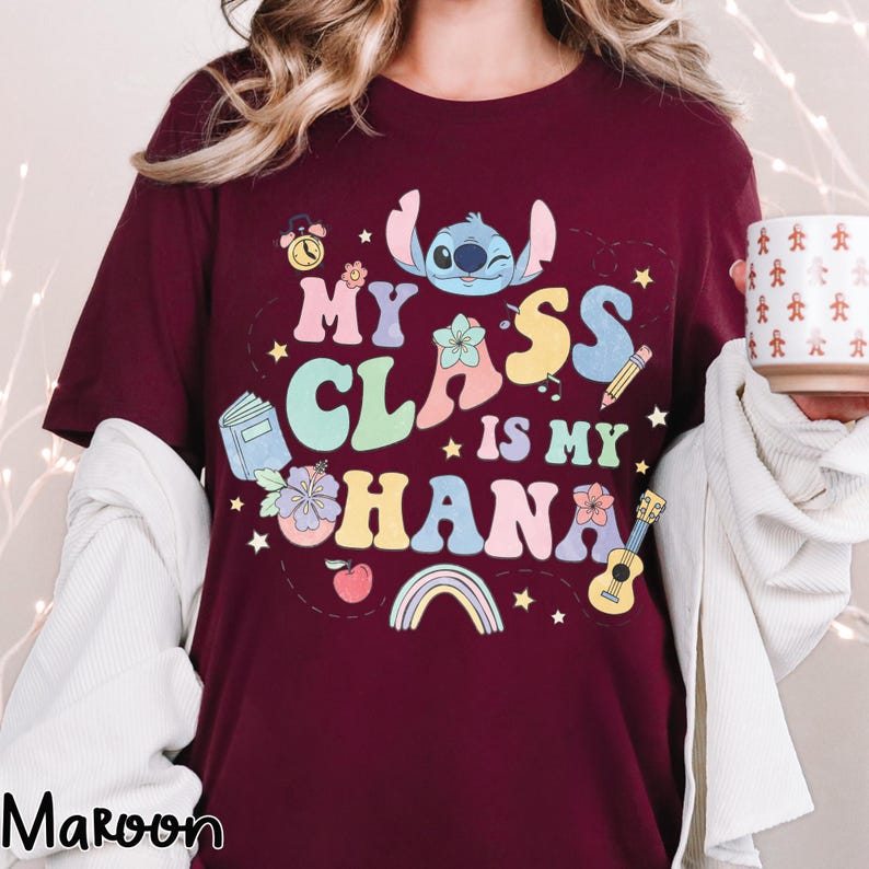 Puede incluir: Camiseta color granate con el texto "My Class is My Ohana" y un personaje de dibujos animados. El dise&ntilde;o incluye un reloj, un libro, flores, un l&aacute;piz, un ukelele y un arco&iacute;ris. La camiseta es de cuello redondo y manga corta.