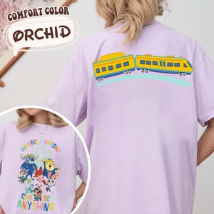 Pu&ograve; includere: T-shirt Orchid Comfort Colors lilla chiaro con un disegno di un treno a fumetti sul retro. La parte anteriore della maglietta presenta una grafica con la scritta "Where Anyone Can Be Anything".