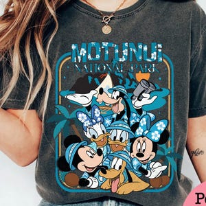 May include: Dark gray t-shirt featuring a graphic of Disney characters, including Mickey, Minnie, Donald, Daisy, Goofy, and Pluto, in Hawaiian shirts and hats. The design includes the text "MOTUNUI NATIONAL PARK" in a blue and white font, with palm trees and a mountain in the background.