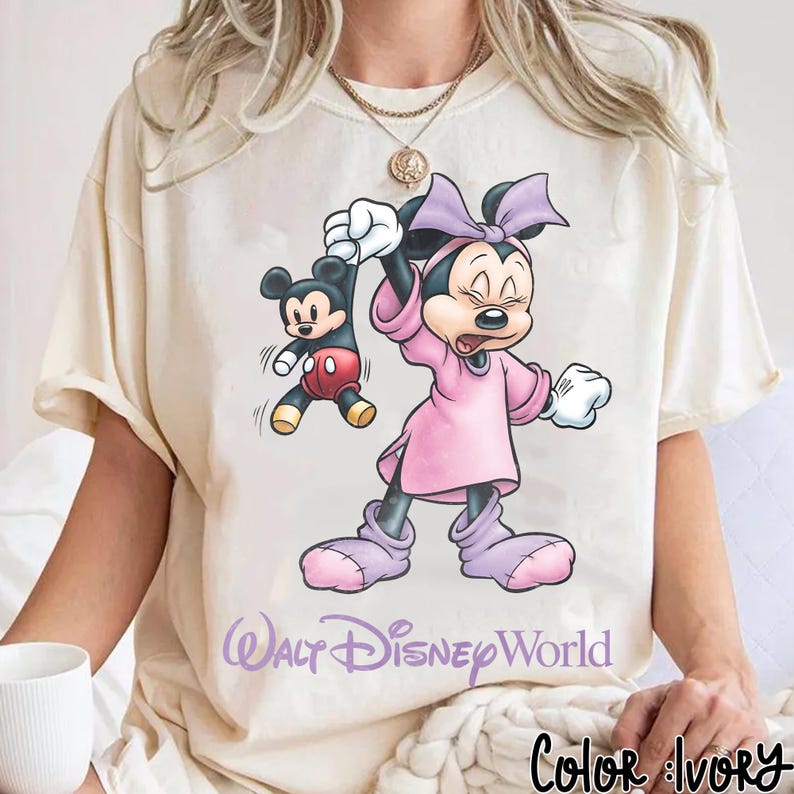 May include: Ivory-colored t-shirt featuring a Minnie Mouse graphic holding a Mickey Mouse figure. Minnie is wearing a pink dress and bow. The text "Walt Disney World" is printed below the graphic. The words "Color: Ivory" are printed at the bottom.