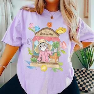 Op de afbeelding: Lichtpaars T-shirt met een cartoonkonijn met een hoed en geruit overhemd, met wortels en groenten in een tuin. Het ontwerp bevat de tekst "2026 Earth Day" en bloemen. Er is ook een gele hoed met "2026".