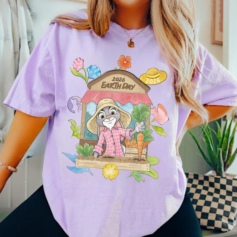 Op de afbeelding: Lichtpaars T-shirt met een cartoonkonijn met een hoed en geruit overhemd, met wortels en groenten in een tuin. Het ontwerp bevat de tekst "2026 Earth Day" en bloemen. Er is ook een gele hoed met "2026".