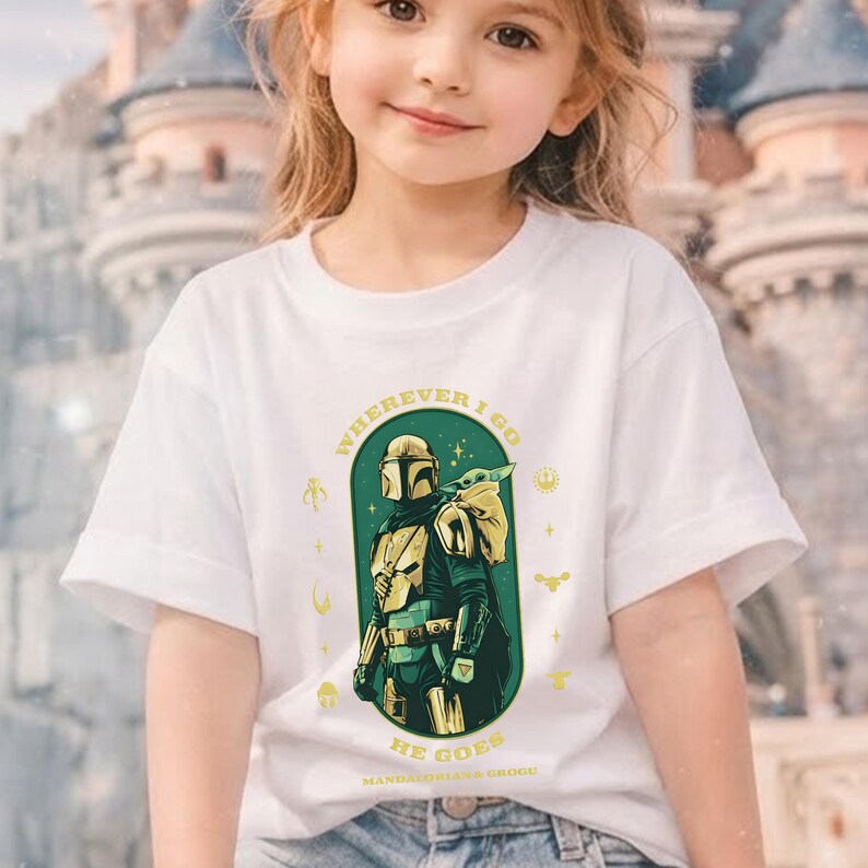 Peut inclure: T-shirt blanc avec un motif de The Mandalorian et Grogu, avec le texte "WHEREVER I GO HE GOES". Le design utilise des tons de vert, d'or et de noir. Le t-shirt est de style col rond.