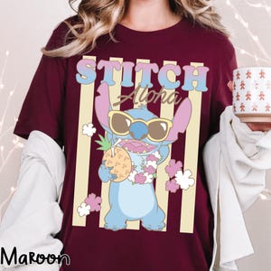 K&ouml;nnte beinhalten: Burgunderfarbenes T-Shirt mit einer Cartoon-Figur von Stitch, die eine Sonnenbrille tr&auml;gt und eine Ananas h&auml;lt. Der Text "STITCH Aloha" befindet sich &uuml;ber der Figur, mit vertikalen gelben Streifen im Hintergrund. Das Wort "Maroon" steht unten.
