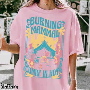 Puede incluir: Camiseta rosa claro con un dise&ntilde;o gr&aacute;fico turquesa y amarillo. El dise&ntilde;o presenta el texto "BURNING MAMMAL COMIN' IN HOT!", una tienda de campa&ntilde;a, un cami&oacute;n de comida y figuras de dibujos animados.