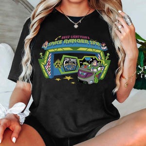 K&ouml;nnte beinhalten: Schwarzes T-Shirt mit einem farbenfrohen Aufdruck von Buzz Lightyear's Space Ranger Spin. Das Design zeigt den Eingang der Attraktion, ein Fahrzeug mit Figuren und den Text "Buzz Lightyear's Space Ranger Spin" und "Star Command".