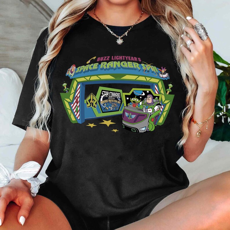K&ouml;nnte beinhalten: Schwarzes T-Shirt mit einem farbenfrohen Aufdruck von Buzz Lightyear's Space Ranger Spin. Das Design zeigt den Eingang der Attraktion, ein Fahrzeug mit Figuren und den Text "Buzz Lightyear's Space Ranger Spin" und "Star Command".