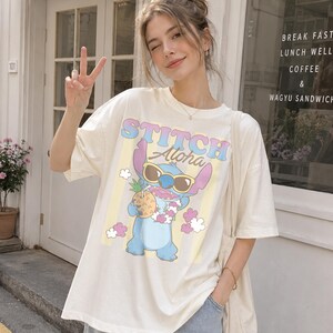 K&ouml;nnte beinhalten: Cremefarbenes T-Shirt mit einem Cartoon-Stitch-Charakter, der eine Sonnenbrille tr&auml;gt und eine Ananas h&auml;lt. Das Shirt hat die Worte "STITCH Aloha" in farbigem Text. Das Shirt ist kurz&auml;rmlig.