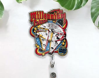 May include: A silver-colored badge reel with a colorful image of the Millennium Falcon spaceship from Star Wars. The image has the text "The Millennium Falcon" and "Light Speed" in a retro style.