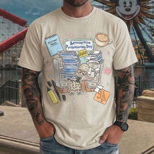 Puede incluir: Camiseta beige con una ilustraci&oacute;n de dibujos animados que celebra el D&iacute;a de los Profesionales Administrativos 2026. El dise&ntilde;o incluye una pila de libros, una taza de caf&eacute; y el texto "Administrative Professionals Day 2026".