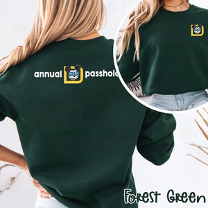 Puede incluir: Sudadera verde bosque con el texto "annual passhold" y un gr&aacute;fico de Mickey Mouse. La sudadera tiene cuello redondo y mangas largas. El gr&aacute;fico es un cuadrado amarillo con una cabeza de Mickey Mouse en su interior.