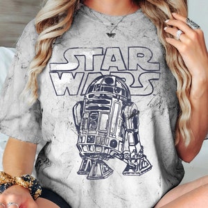 May include: A white t-shirt with a distressed look featuring a black and white graphic of R2-D2 from Star Wars. The text "Star Wars" is printed in black above the robot.