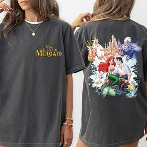 May include: A dark gray t-shirt with a graphic print of Ariel, Prince Eric, and Ursula from Disney's The Little Mermaid. The text "Disney The Little Mermaid" is printed on the front of the shirt.