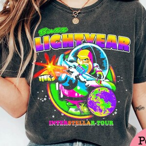 May include: Dark gray t-shirt featuring a colorful graphic of Buzz Lightyear in a space suit, firing a laser. The text "Buzz Lightyear" is above the image, and "Interstellar Tour" is below.
