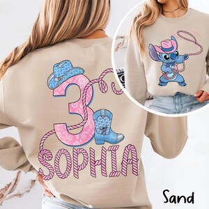 May include: Sand-colored sweatshirt featuring a graphic of a cartoon character wearing a cowboy hat and holding a lasso. The back of the sweatshirt has a pink and blue design with the number 3, a cowboy boot, a cowboy hat, and the name "Sophia".