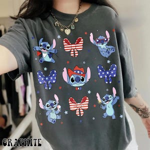 May include: Graphite-coloured t-shirt with a design of cartoon Stitch characters in various poses, alongside patriotic bow ties and accessories. The design includes red, white, and blue stars and bow tie graphics.