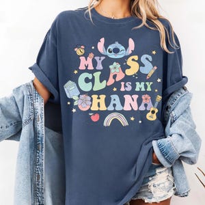 Puede incluir: Camiseta azul marino con el texto "My Class is My Ohana" en letras coloridas, con un personaje de dibujos animados, flores y gr&aacute;ficos escolares. La camiseta tiene las mangas dobladas.