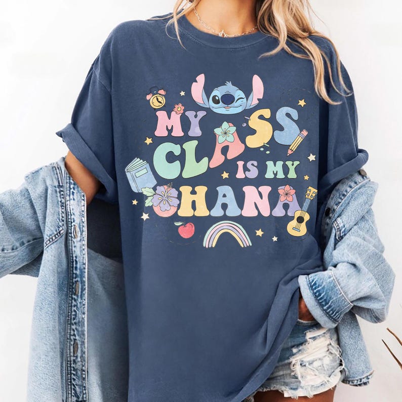 Puede incluir: Camiseta azul marino con el texto "My Class is My Ohana" en letras coloridas, con un personaje de dibujos animados, flores y gr&aacute;ficos escolares. La camiseta tiene las mangas dobladas.