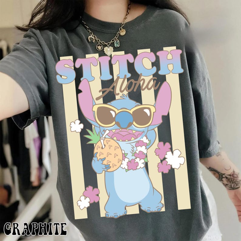 K&ouml;nnte beinhalten: Graphitgraues T-Shirt mit einer Cartoon-Figur von Stitch, die eine Sonnenbrille tr&auml;gt und eine Ananas h&auml;lt. Das Shirt hat die Worte "STITCH Aloha" in einer bunten Schrift, mit vertikalen beigen Streifen und wei&szlig;en Blumenakzenten.