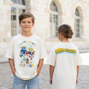 Pu&ograve; includere: Due bambini indossano magliette color crema. La parte anteriore della prima maglietta presenta una grafica colorata con la scritta "WHERE ANYONE CAN BE ANYTHING". Il retro della seconda maglietta presenta un disegno di treno giallo e blu.