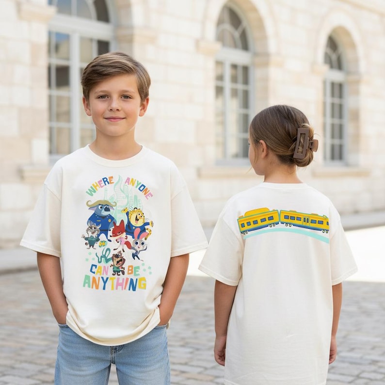 Pu&ograve; includere: Due bambini indossano magliette color crema. La parte anteriore della prima maglietta presenta una grafica colorata con la scritta "WHERE ANYONE CAN BE ANYTHING". Il retro della seconda maglietta presenta un disegno di treno giallo e blu.