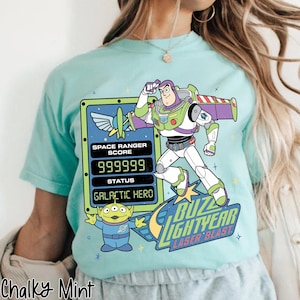 May include: A light blue t-shirt featuring a graphic of Buzz Lightyear from Toy Story. The design includes Buzz Lightyear, an alien, and text that reads "Buzz Lightyear Laser Blast". The shirt also has a "Space Ranger Score" graphic.