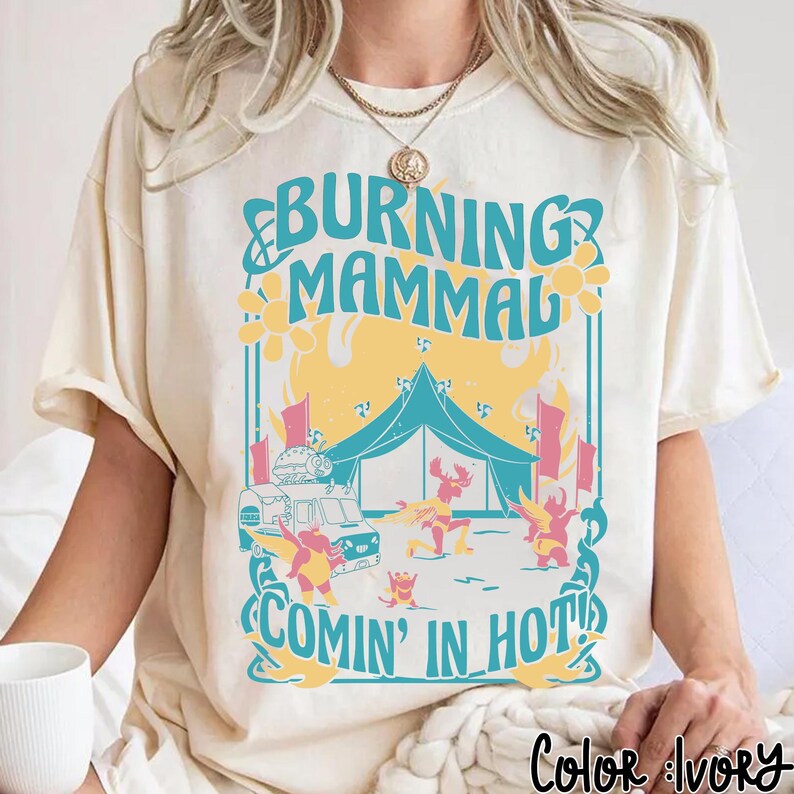 Puede incluir: Camiseta de color marfil con un dise&ntilde;o gr&aacute;fico. El dise&ntilde;o presenta el texto "BURNING MAMMAL COMIN' IN HOT!" en turquesa y amarillo. El dise&ntilde;o incluye una tienda de campa&ntilde;a, un cami&oacute;n de comida y figuras de dibujos animados.