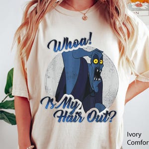 Puede incluir: Camiseta de color marfil con un gr&aacute;fico desgastado de un personaje de dibujos animados azul con ojos amarillos. El texto "Whoa! Is My Hair Out?" est&aacute; impreso encima y debajo del personaje en una fuente azul. La camiseta es de la marca Comfort Color.
