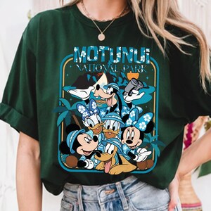 May include: Dark green t-shirt featuring a graphic of Disney characters, including Mickey, Minnie, Donald, Daisy, Goofy, and Pluto, in Hawaiian shirts and hats. The shirt has the text "MOTUNUI NATIONAL PARK" in a blue and white design.